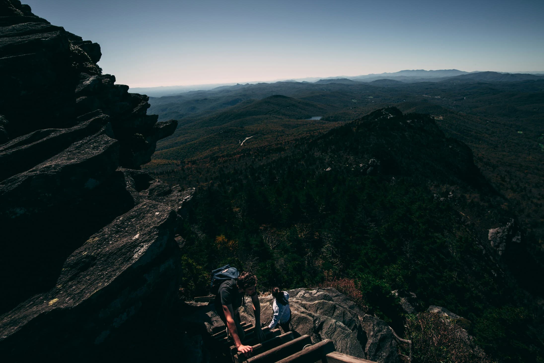 Two hikers climbing wooden steps toward a rocky mountain summit overlooking a vast forested ridgeline